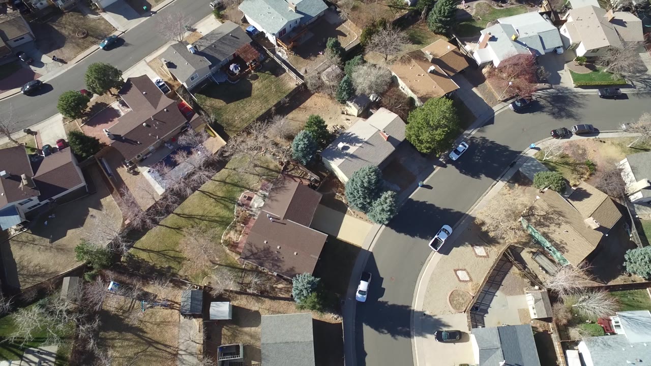 Aerial frame over Hidden Lake, Colorado showing brown and light rooftops, fenced backyards, a cul-de-sac-like curve, parked vehicles, and strong tree shadows across lawns.