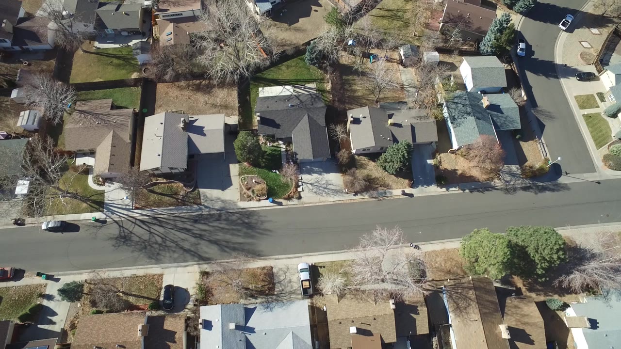 Aerial frame over Hidden Lake, Colorado showing a residential street crossing the center, rows of rooftops, fenced yards, trees casting shadows, and no visible people.