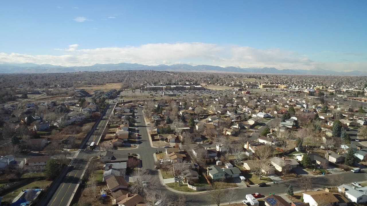 Aerial frame in Hidden Lake, Colorado showing a broad residential grid, a main road at left, scattered trees, open ground, and mountains in the distance, with no visible people.