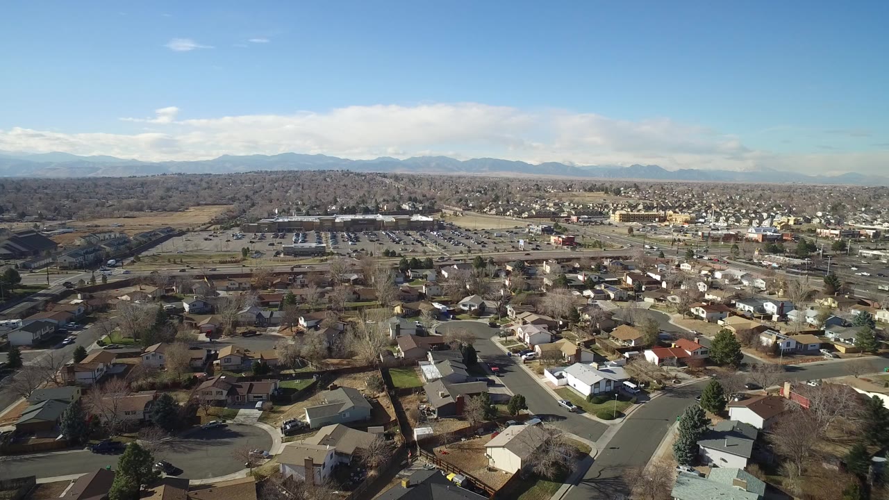 Opening aerial frame over Hidden Lake, Colorado with foreground rooftops, curving residential streets, parking lots beyond, a mountain horizon, and no visible people.