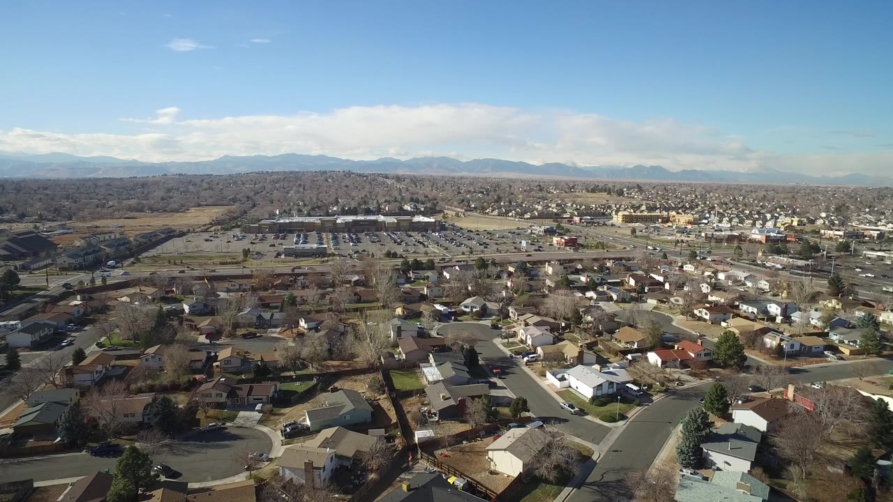 Second aerial frame in Hidden Lake, Colorado showing residential blocks below, a broad parking area in the midground, low buildings, distant mountains, and no visible people.