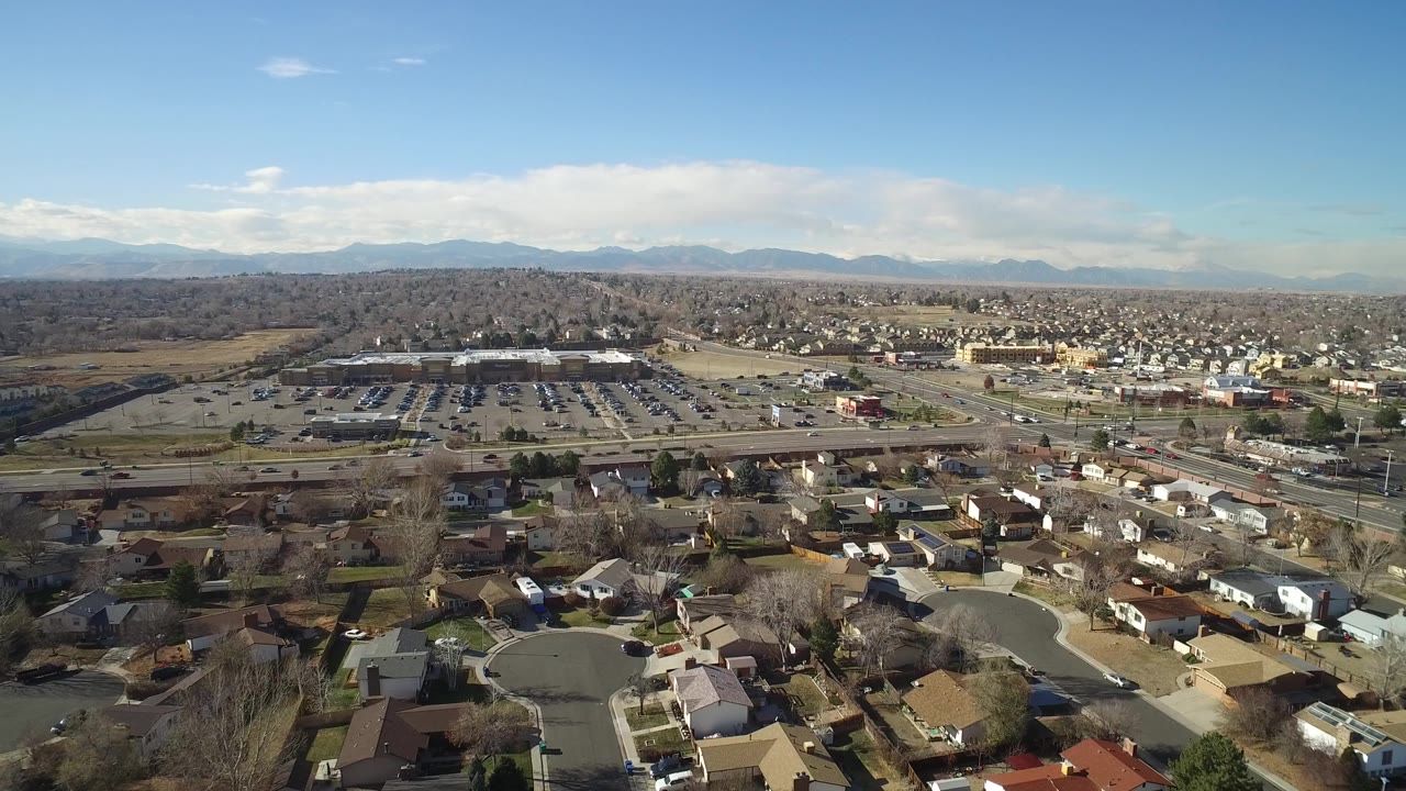 Aerial frame in Hidden Lake, Colorado showing more of the residential blocks at lower frame, a main road with small cars, large parking areas, distant tree cover, mountains, and no visible people.