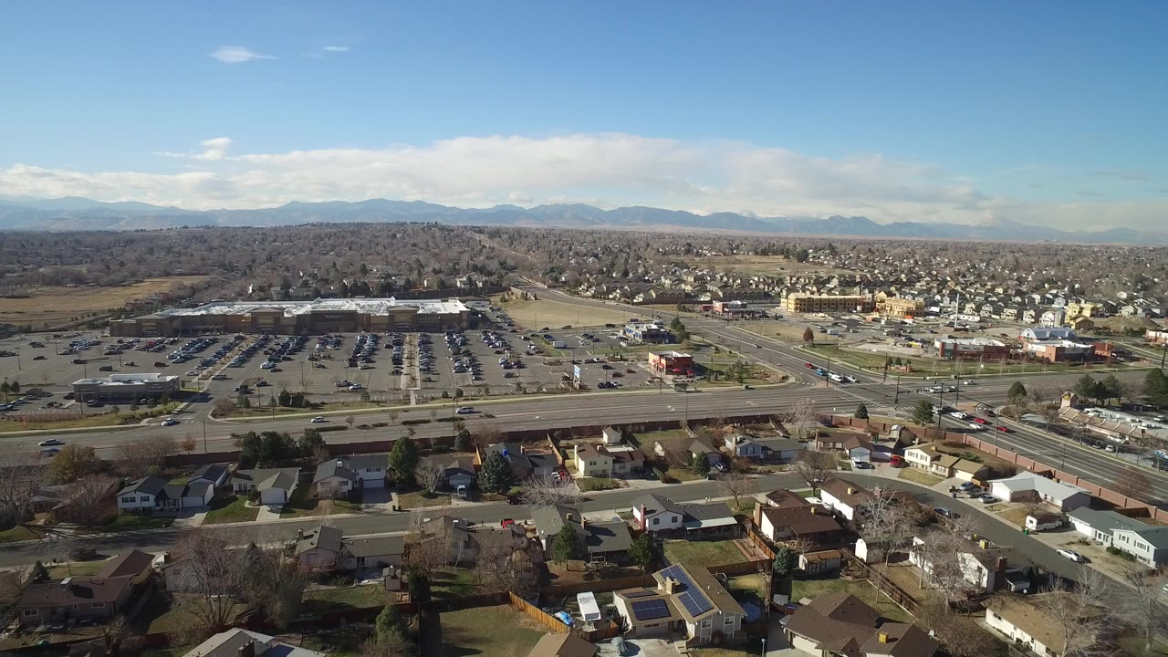 Aerial view of suburban homes, a broad road, parking lot, and distant hills under blue sky.