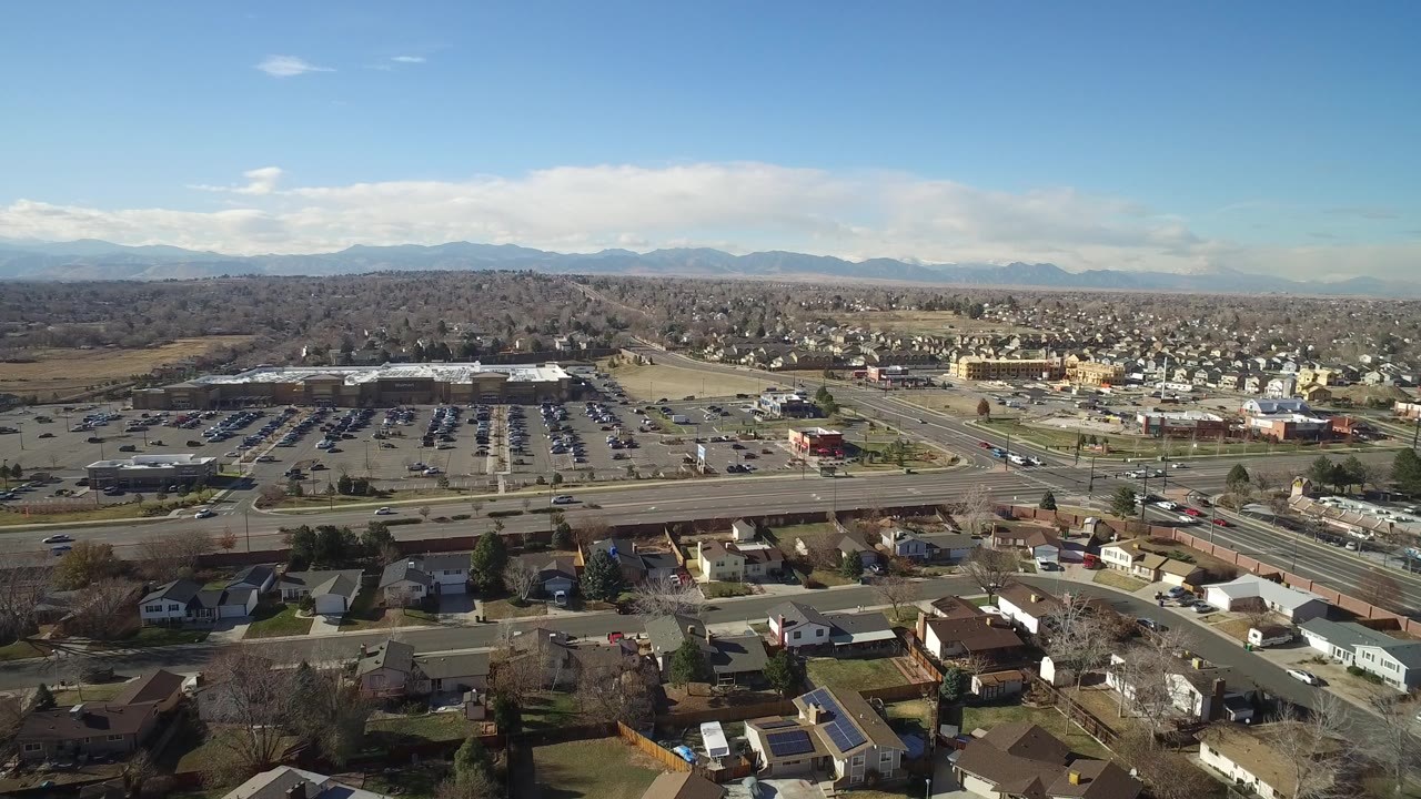 Early aerial view in Hidden Lake, Colorado, with rooftops along the lower edge, a busy road corridor, a large parking area, scattered commercial buildings, distant mountains, and no visible people.