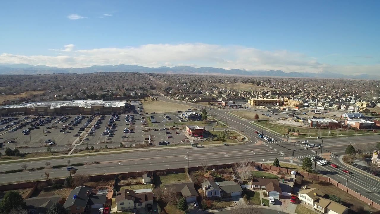 Aerial view of a wide road, parking lots, buildings, homes, and distant hills.