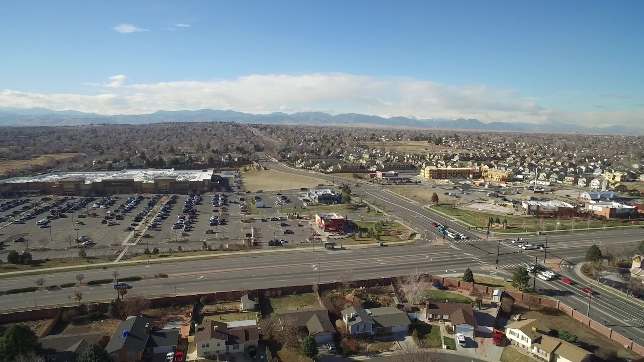 A slightly later aerial view in Hidden Lake, Colorado centers the parking lots and arterial road, with houses, low buildings, distant hills, clear sky, and no visible people.