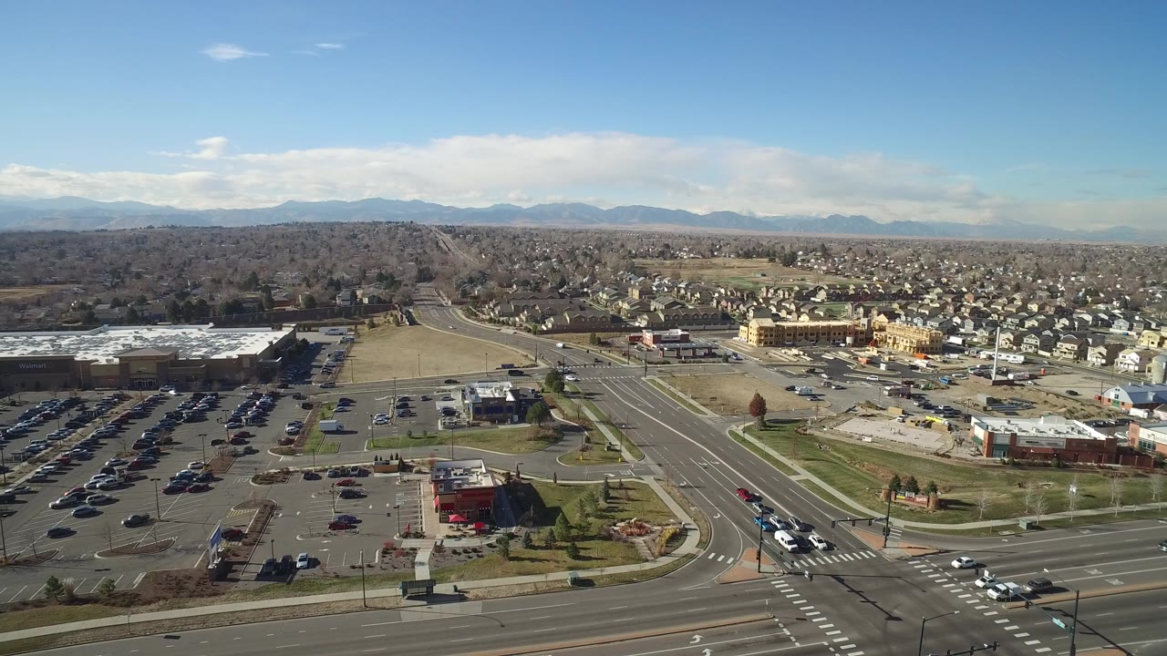 Elevated daylight view over Hidden Lake, Colorado, with a multi-lane road in front, parking lots and businesses in the middle distance, homes and mountains beyond, and no visible people.