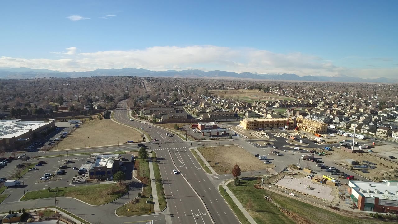 High aerial frame over Hidden Lake, Colorado, showing a broad road corridor, parking lots, rows of homes, leafless trees, distant mountains, and no visible people.