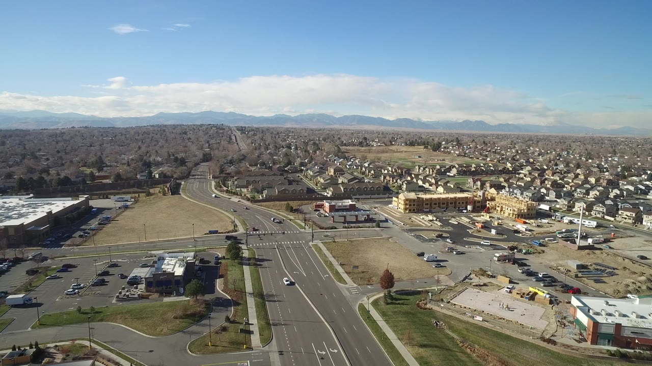 Aerial frame in Hidden Lake, Colorado, with the intersection centered lower in the view, commercial roofs and lots nearby, dense housing beyond, mountains on the horizon, and no visible people.