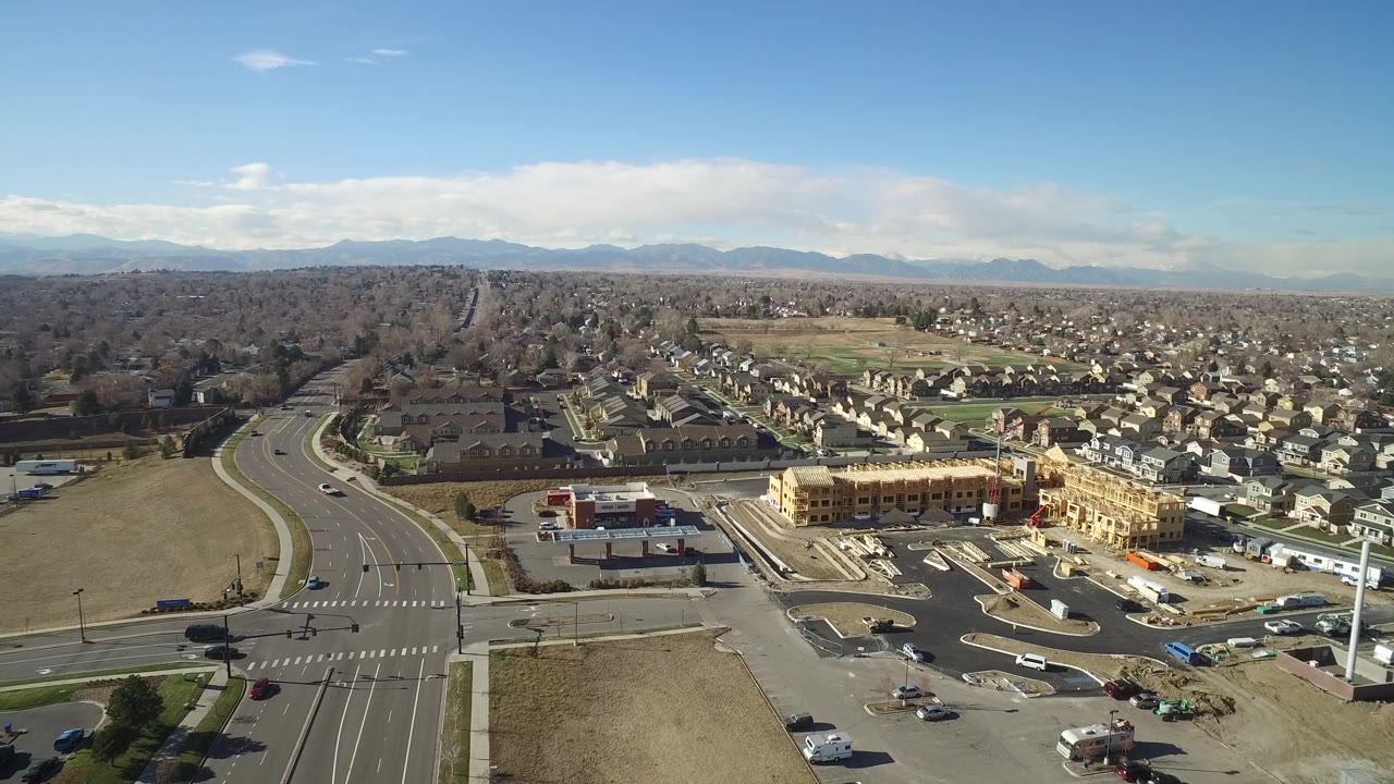 Aerial frame in Hidden Lake, Colorado with the curving road and intersection lower left, commercial parking areas near center, active building framing at lower right, residential blocks behind, and no visible people.