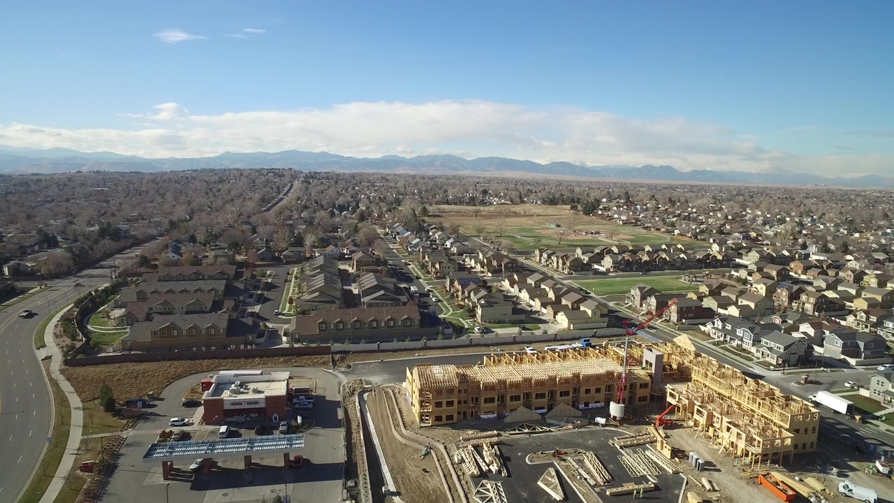 Opening aerial frame over Hidden Lake, Colorado, with a wood-framed construction site at the bottom, suburban homes beyond, distant mountains, blue sky, and no visible people.