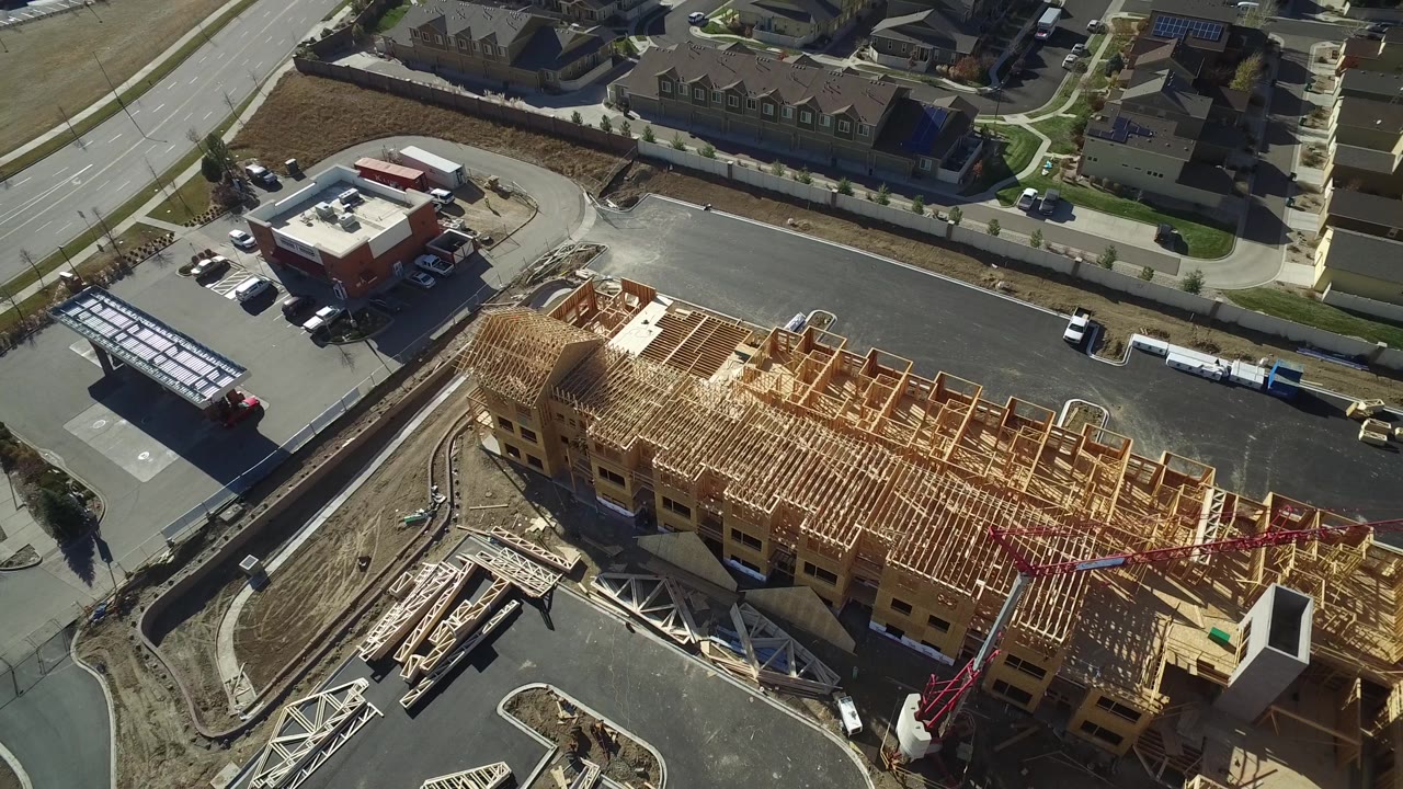 Aerial view in Hidden Lake, Colorado with roof trusses and wall framing filling the lower frame, a parking area and small commercial-looking building above, and no visible people.