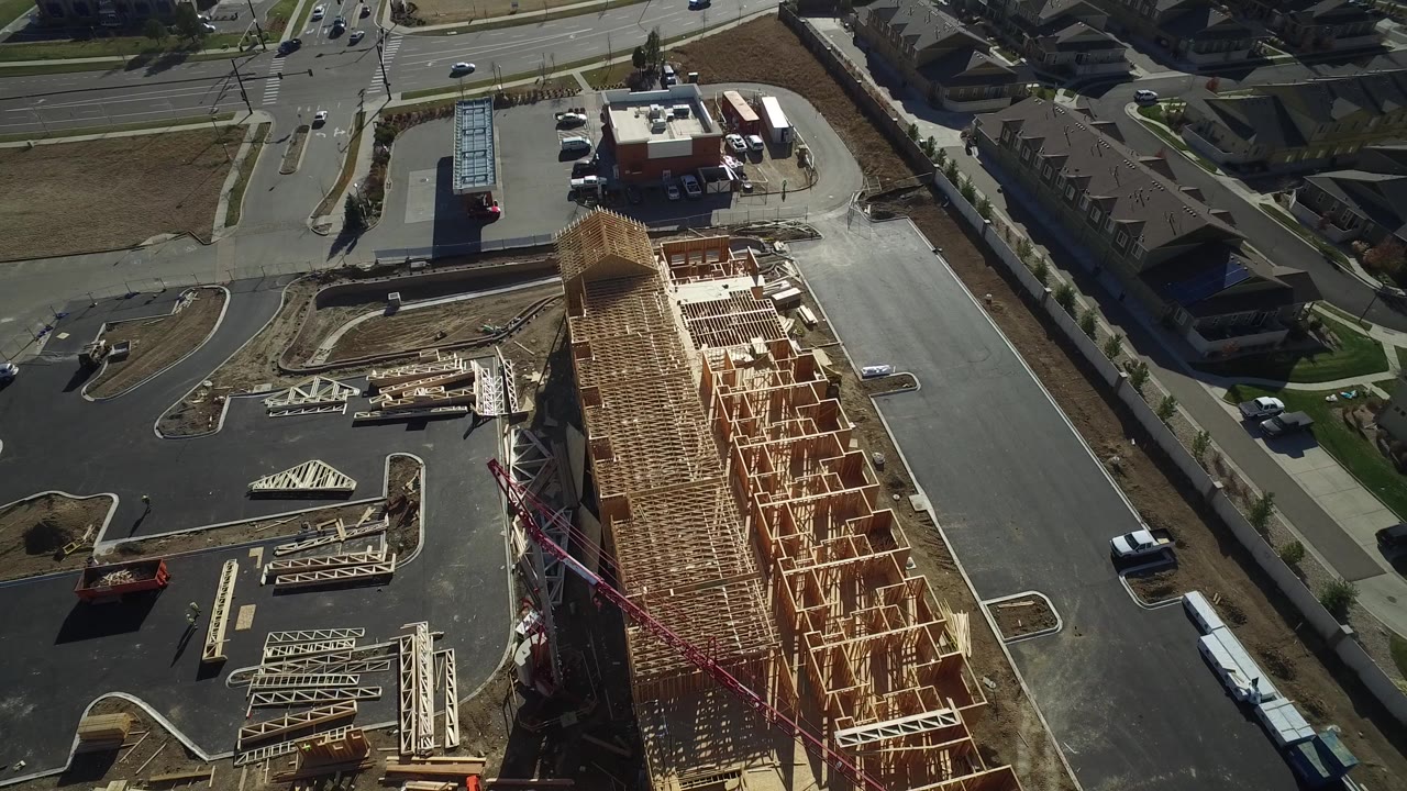 High aerial frame over Hidden Lake, Colorado showing a long wood-framed building under construction beside paved roads, parking lots, nearby houses, and no visible people.