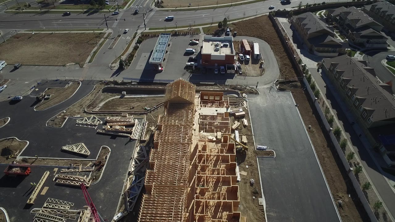 Aerial frame from Hidden Lake, Colorado with exposed roof trusses dominating the foreground, a parking lot and small commercial building nearby, road traffic above, and no visible people.