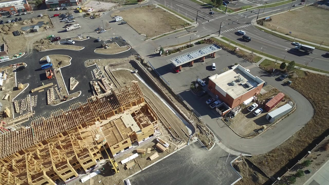 Aerial frame over Hidden Lake, Colorado showing a wood-framed construction site, paved roads, parked vehicles, graded dirt, and a nearby low commercial building with no visible people.
