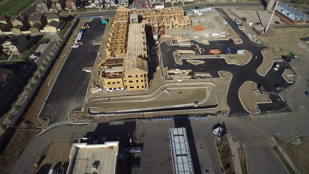 Aerial view in Hidden Lake, Colorado with the wood-framed building centered higher in frame, parking pavement on both sides, scattered vehicles, construction supplies, and no visible people.