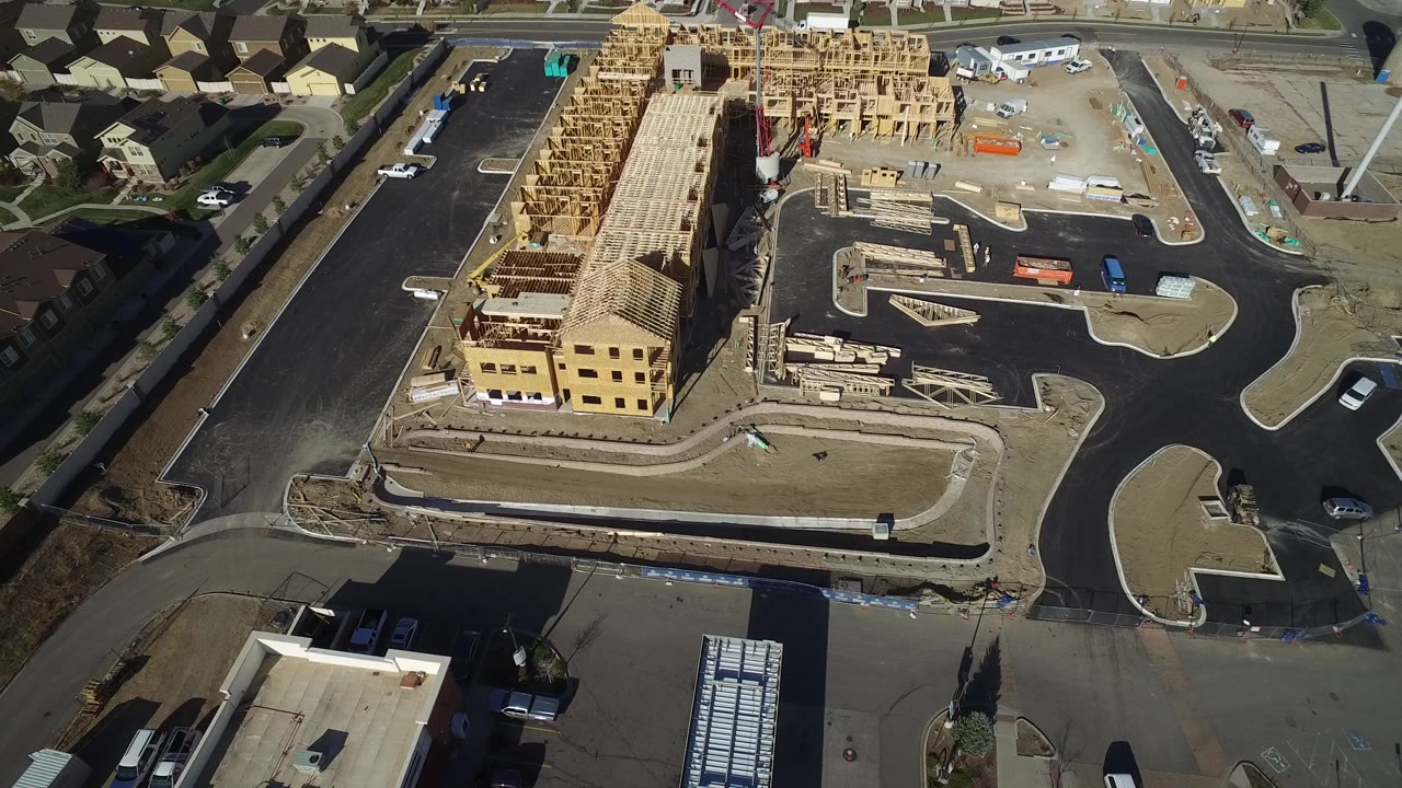 Aerial view in Hidden Lake, Colorado showing a wood-framed multi-unit building under construction, paved roads, curb cuts, nearby homes, and no visible people.