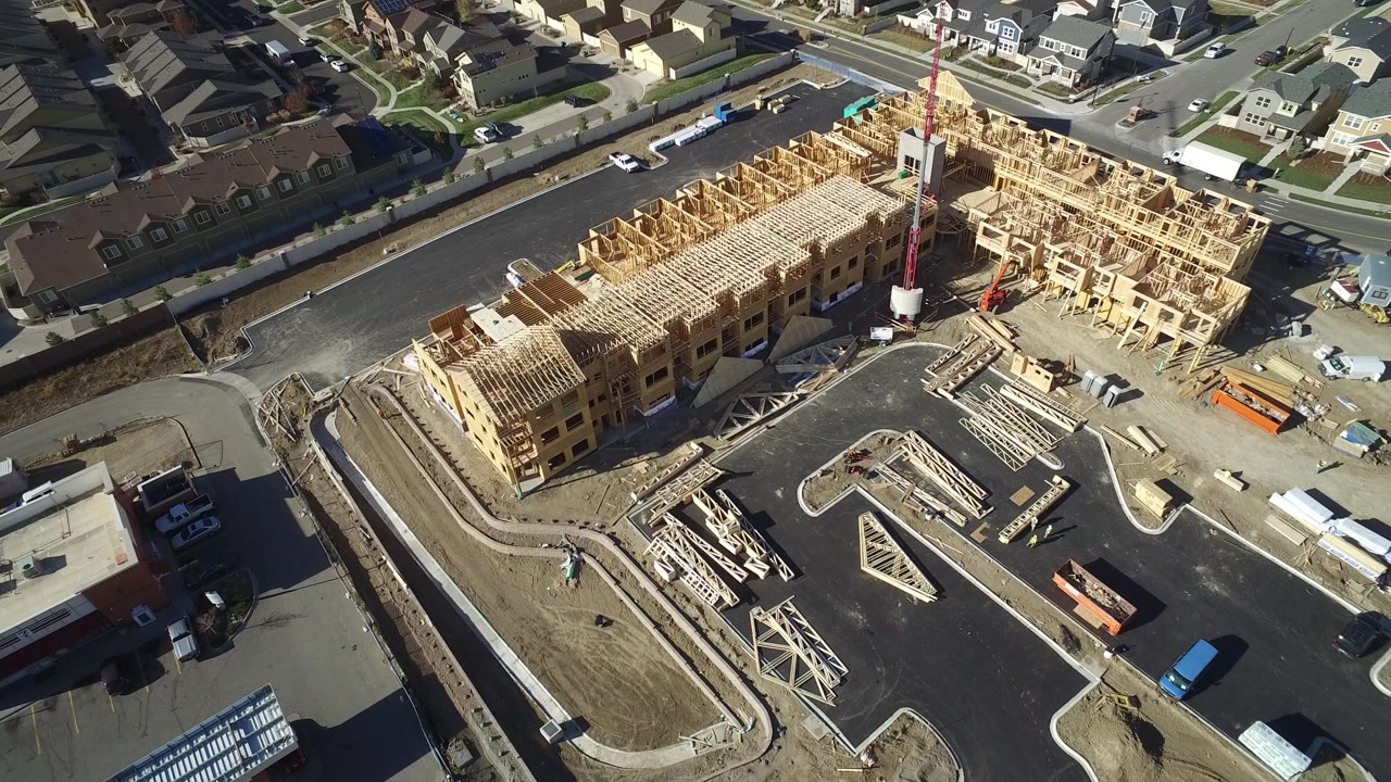 High aerial view in Hidden Lake, Colorado showing bare wood residential framing, paved parking areas, stacked lumber, roads, and surrounding homes, with no visible people.