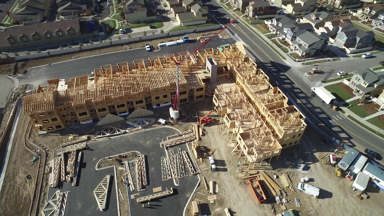 Oblique drone view in Hidden Lake, Colorado showing the framed structure more centered, with a crane or lift near the site, neighboring homes, streets, and no visible people.