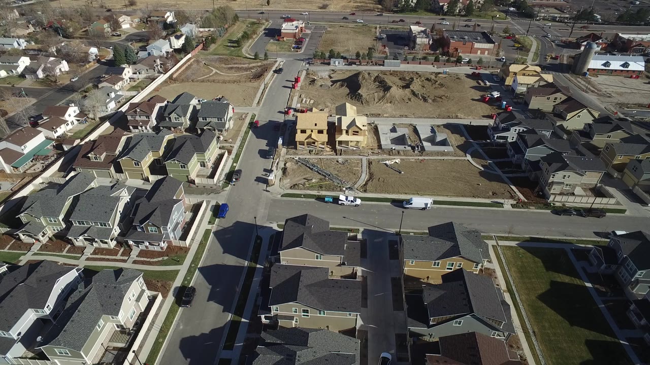 Aerial view in Hidden Lake, Colorado with clustered rooftops in the foreground, a street with parked vans and cars, and a bare lot under construction near the center, no visible people.