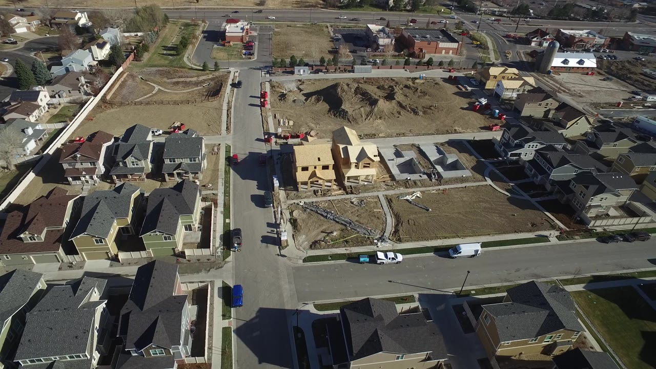 Drone-height view in Hidden Lake, Colorado with a cross street, rooflines, parked cars, bare lots, and a partially framed house, with no visible people.