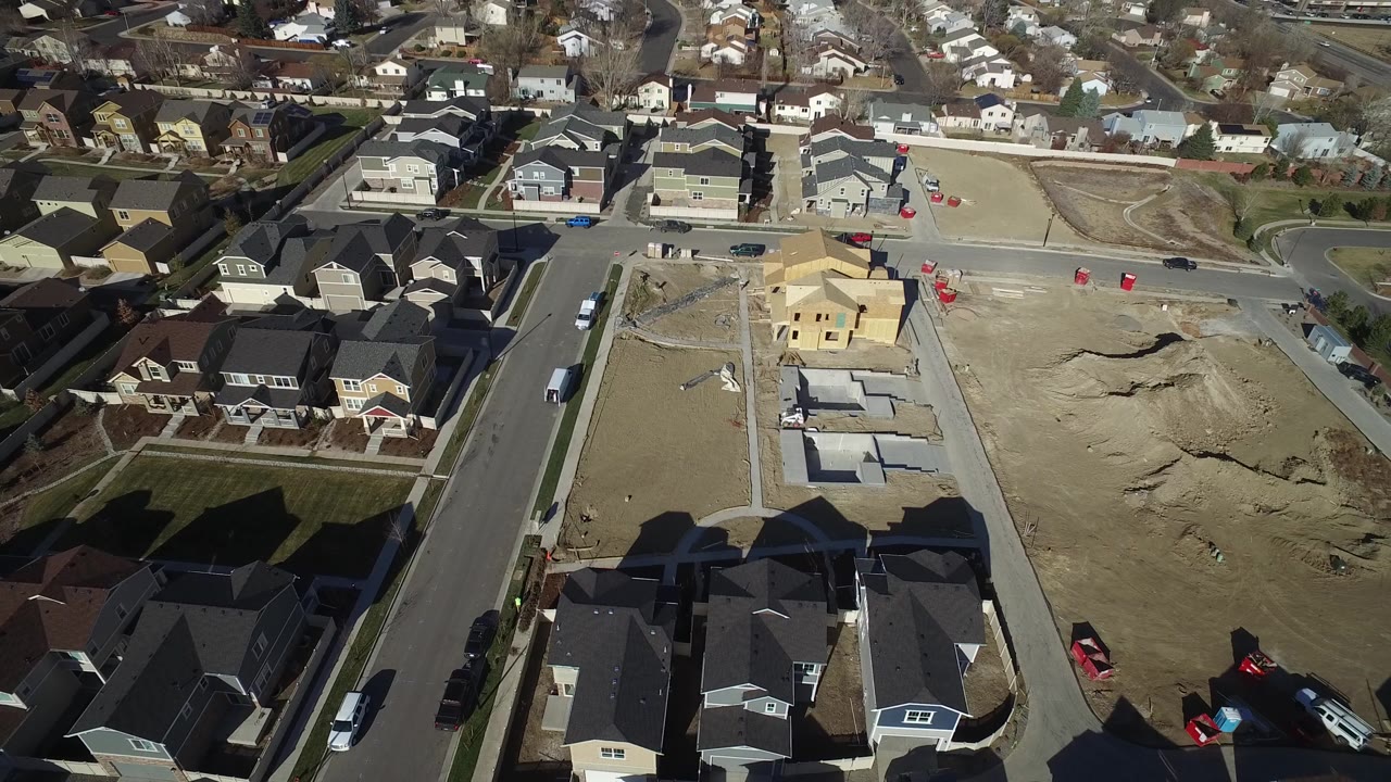 Aerial frame from Hidden Lake, Colorado showing a straight street, completed houses, graded parcels, foundation pads, and a wood-framed home, with no visible people.