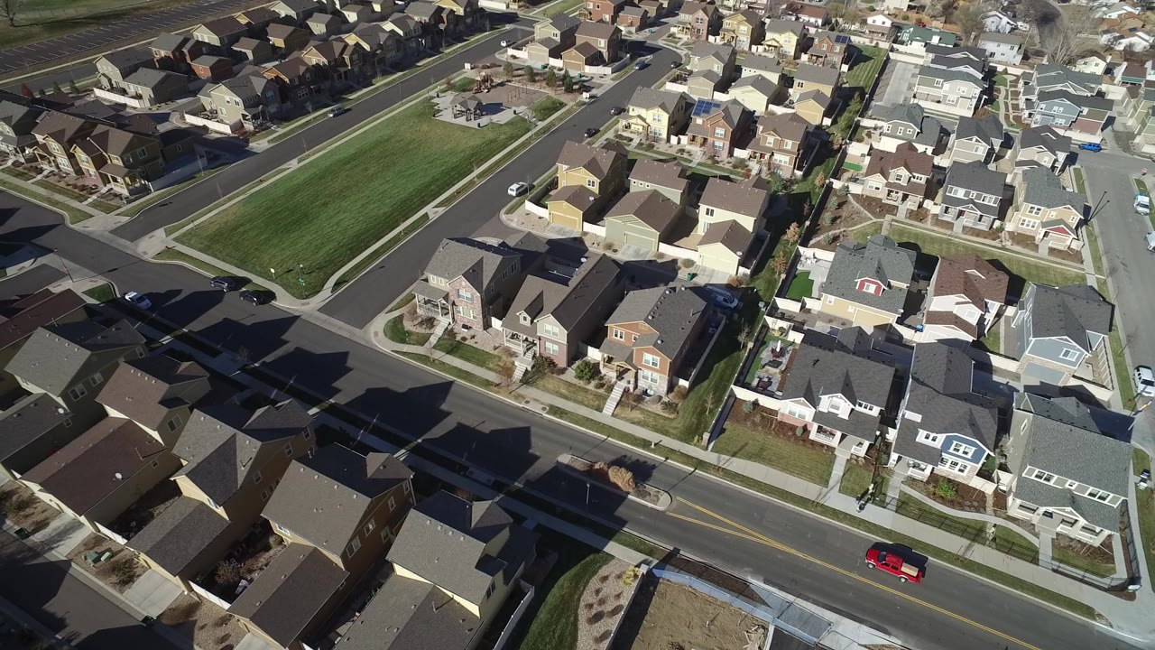 Aerial view of suburban houses, streets, lawns, and a red vehicle on a road.