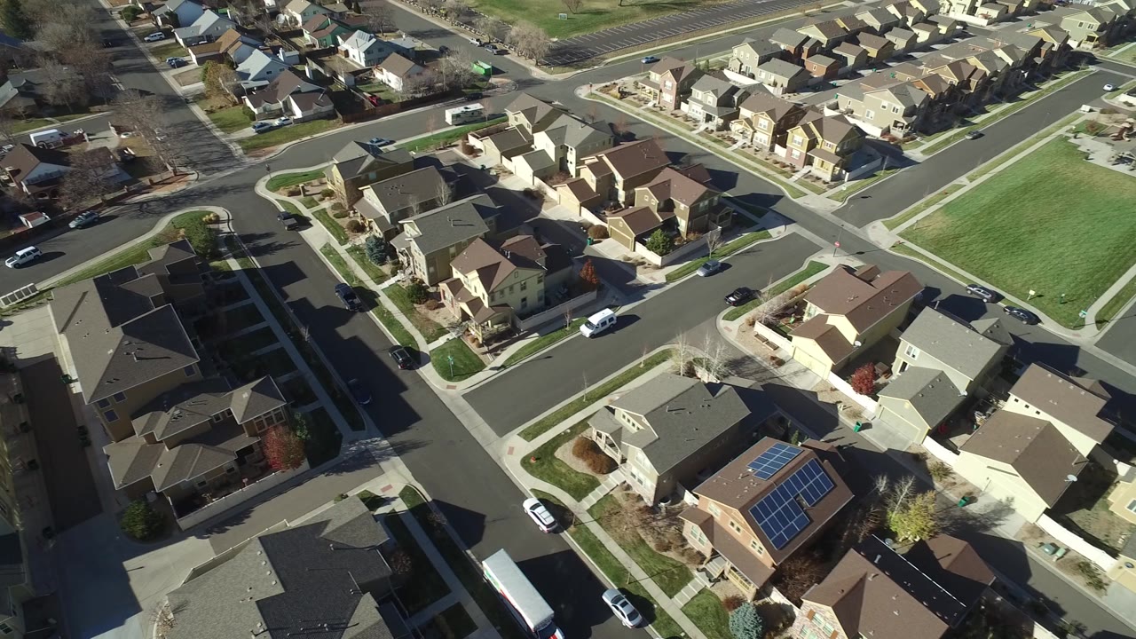 High aerial frame of Hidden Lake, Colorado showing tan houses, gray roofs, a rectangular green field, curving streets, and small vehicles, with no visible people.
