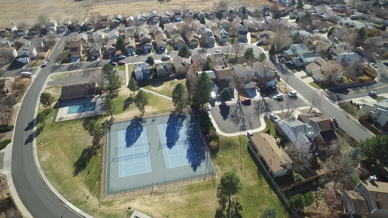 Aerial view of tennis courts, pool, homes, roads, lawns, trees, and parked cars.