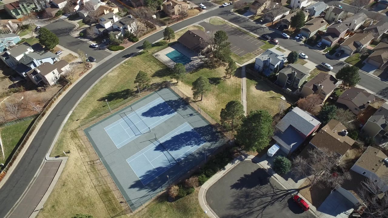 High aerial frame over Hidden Lake, Colorado showing two blue tennis courts, surrounding lawns, nearby single-family houses, parked cars, leafless trees, and no visible people.