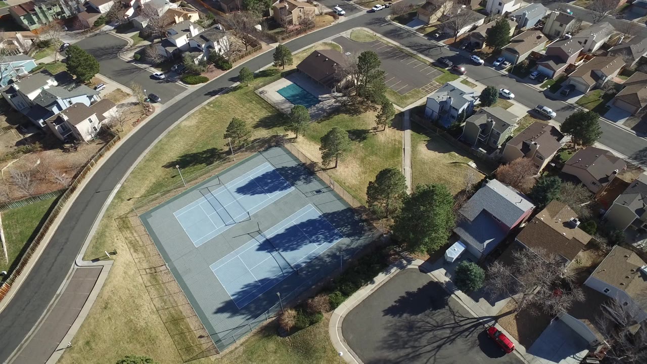 Aerial view in Hidden Lake, Colorado with the tennis courts centered lower in frame, houses and driveways across the top, a curving road at right, and no visible people.