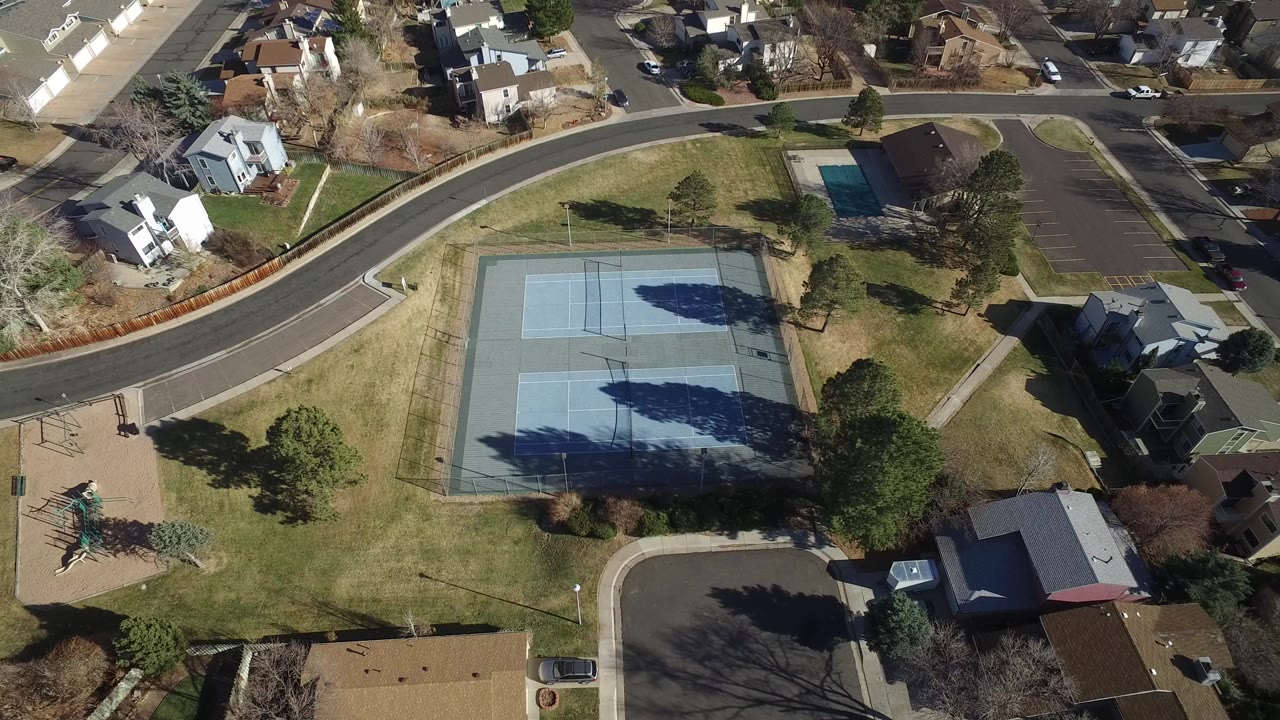 High aerial view in Hidden Lake, Colorado showing blue tennis courts, surrounding grass, curving roads, detached homes, a playground, and no visible people.