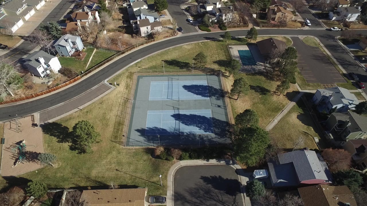 Drone-style view over a park, tennis courts, homes, and parking area.