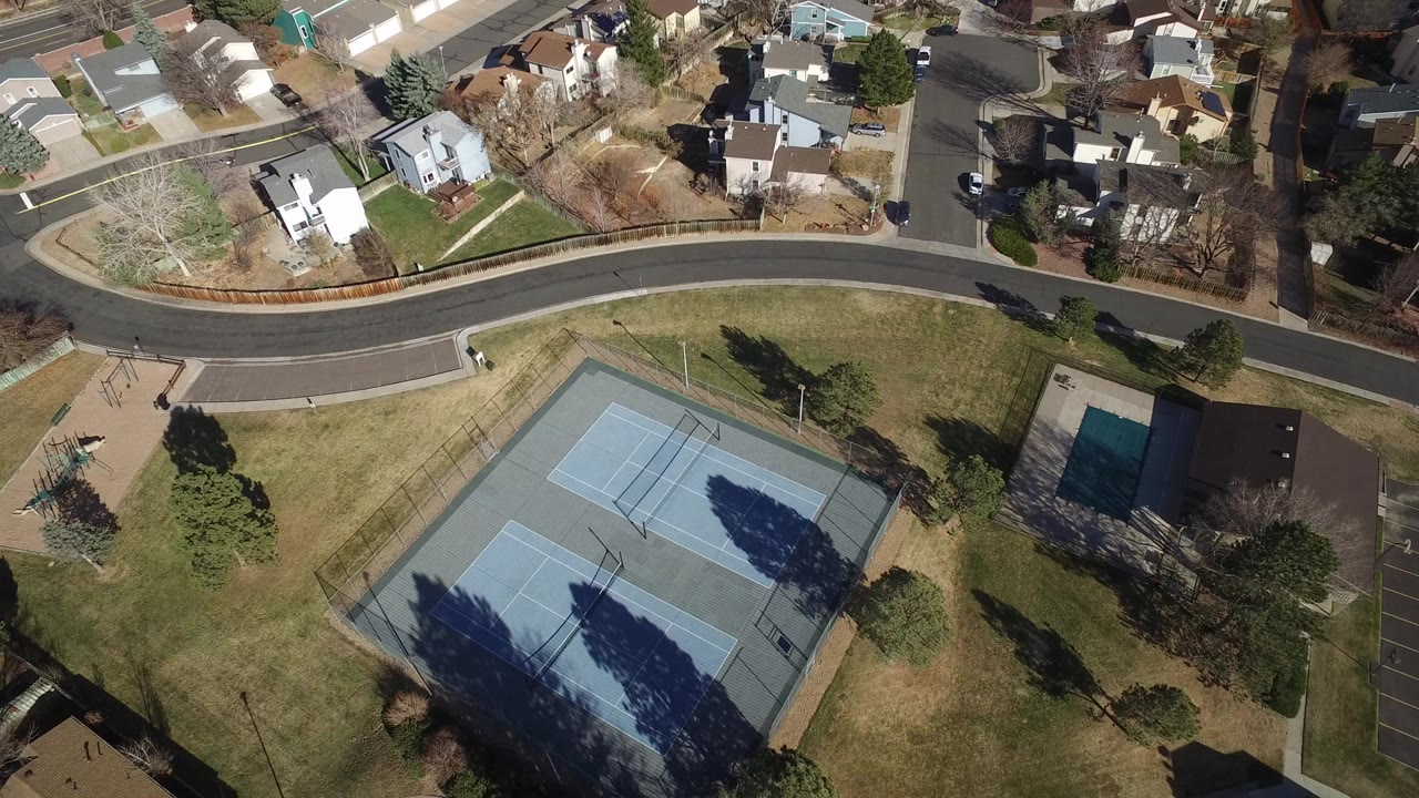 Aerial view of a curved neighborhood road, homes, tennis courts, lawns, and trees.