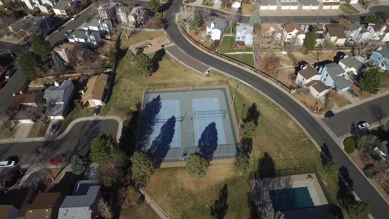 Aerial view in Hidden Lake, Colorado, with the tennis courts slightly lower in frame, bordered by lawns, evergreen trees, residential roads, houses, and no visible people.