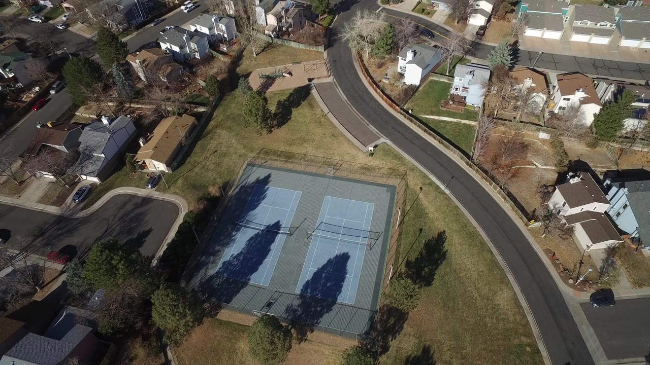 Aerial frame in Hidden Lake, Colorado, with the tennis courts slightly lower in view, a dark tree shadow crossing the court surface, houses around curved streets, and no visible people.