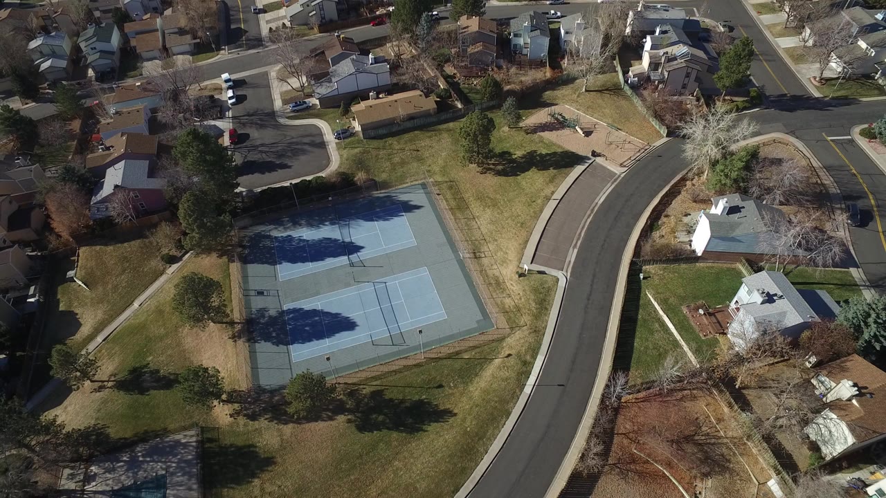 Aerial view of blue tennis courts beside a curved suburban road and homes.
