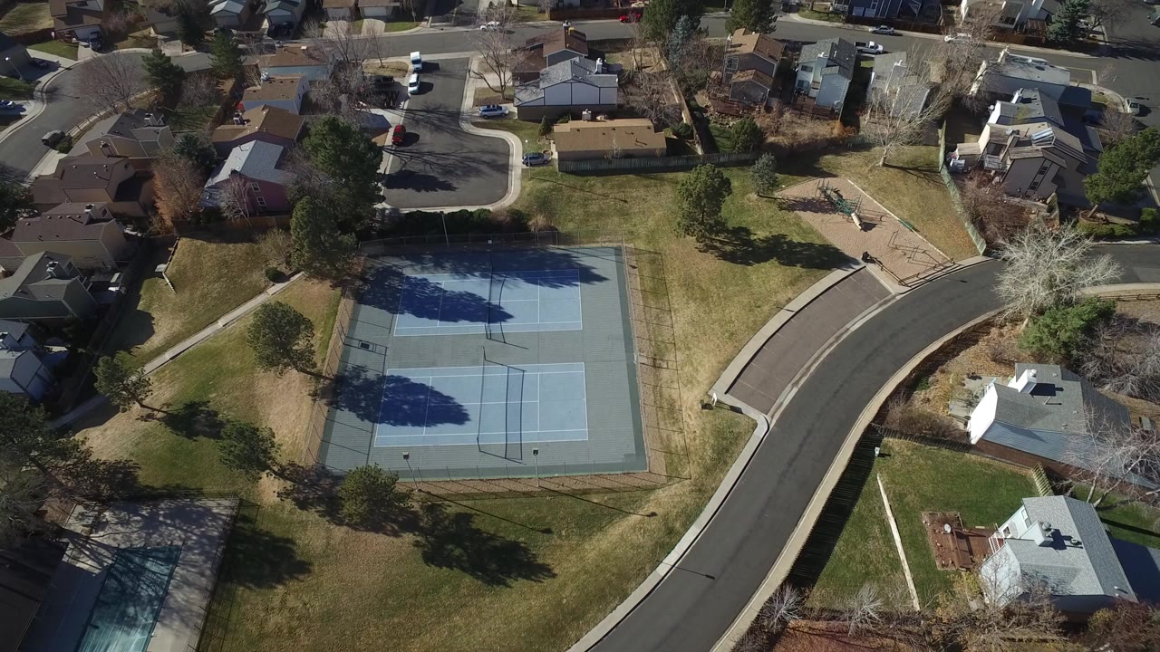 Aerial view of blue tennis courts beside a curved residential road and houses.