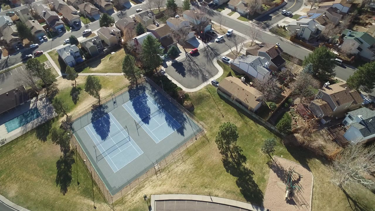 Aerial frame over Hidden Lake, Colorado showing two blue tennis courts, nearby lawns, houses, curving streets, parked cars, and no clearly identifiable people.