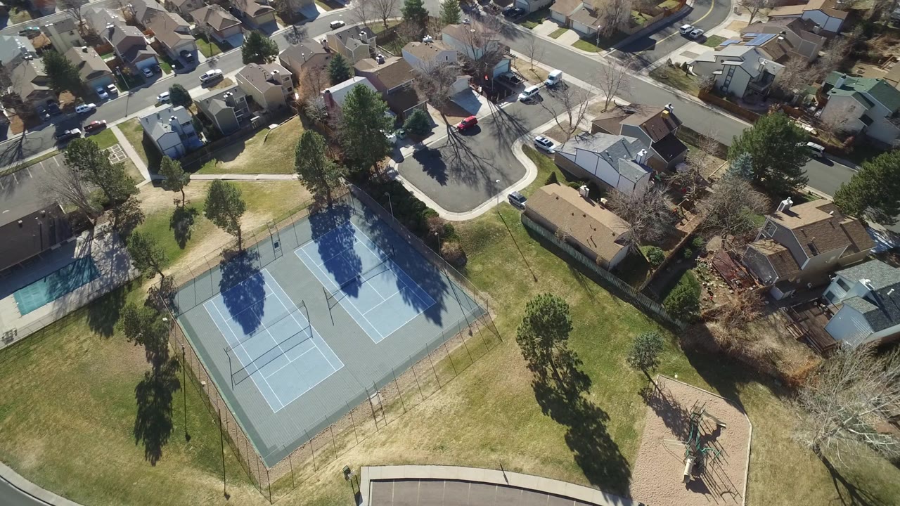 Aerial view in Hidden Lake, Colorado with the twin tennis courts centered lower in frame, open grass around them, detached homes, cars, and a curved residential road.
