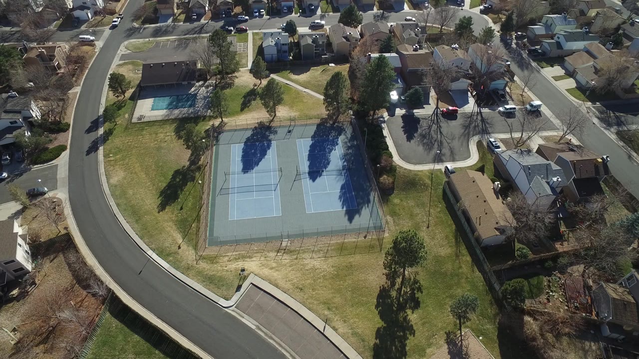 Aerial view in Hidden Lake, Colorado with the two courts centered below homes, a small fenced pool at upper left, long tree shadows, and no visible people.