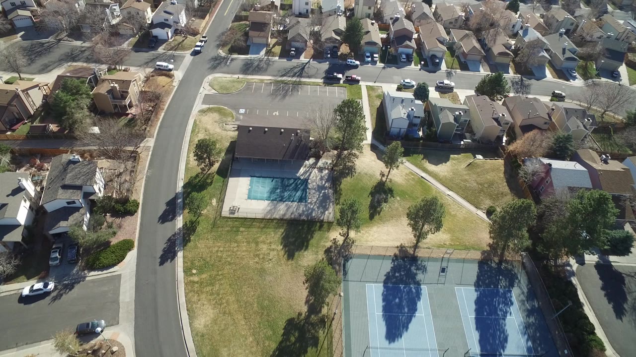 Overhead frame in Hidden Lake, Colorado showing the pool building, blue tennis courts, residential streets, roofs, parked cars, trees, and no clearly visible people.