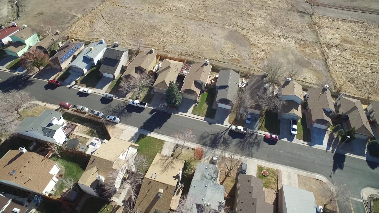 A bird's-eye view in Hidden Lake, Colorado frames tan and gray rooftops, a quiet street with parked vehicles, and open brown land behind the houses, with no visible people.