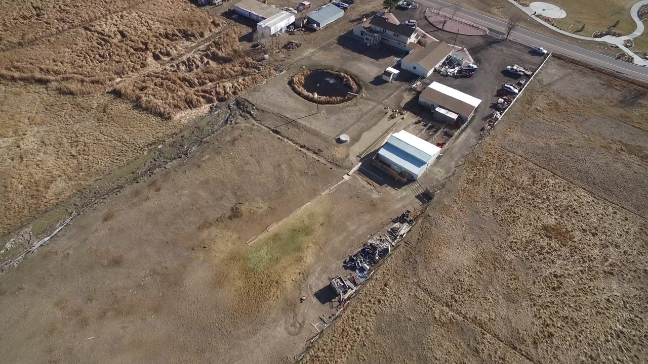 High overhead frame of an industrial yard in Hidden Lake, Colorado, with dry grassland, low buildings, parked vehicles, a road, and no visible people.