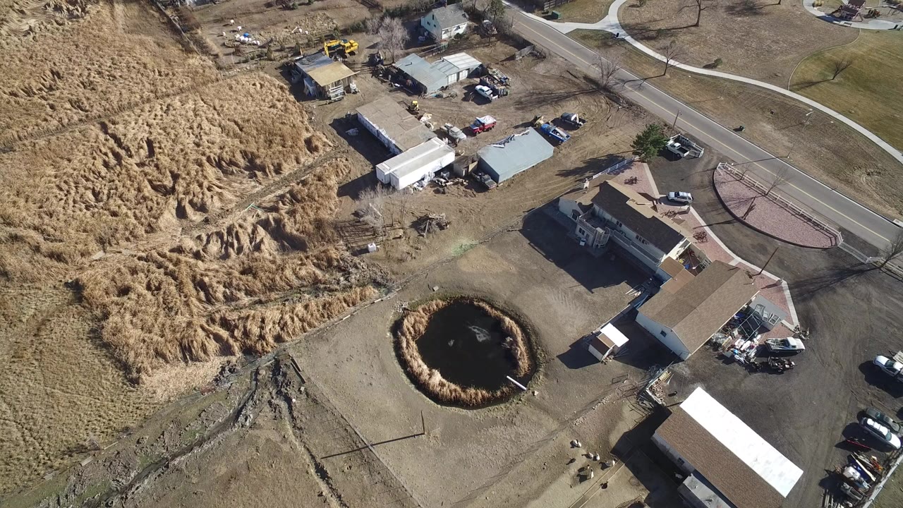 Top-down aerial view of homes, road, dry grass, and a circular pond-like basin.