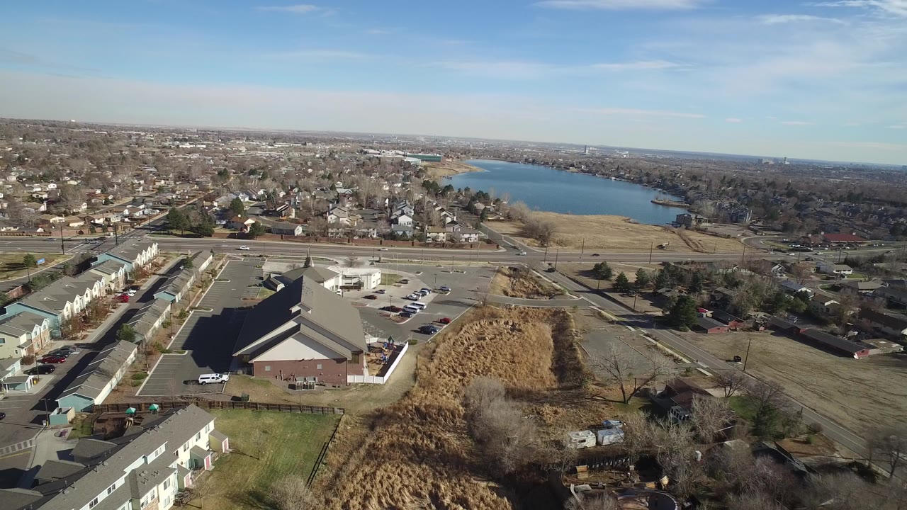 Aerial view of suburban buildings, roads, dry fields, and a lake beyond.