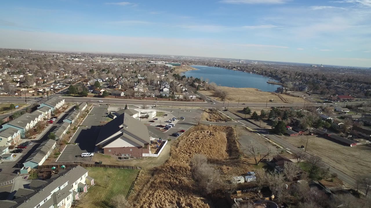 Aerial view in Hidden Lake, Colorado with apartment buildings at left, a large building and parking lot below, road traffic in the midground, the lake in the distance, and no visible people.