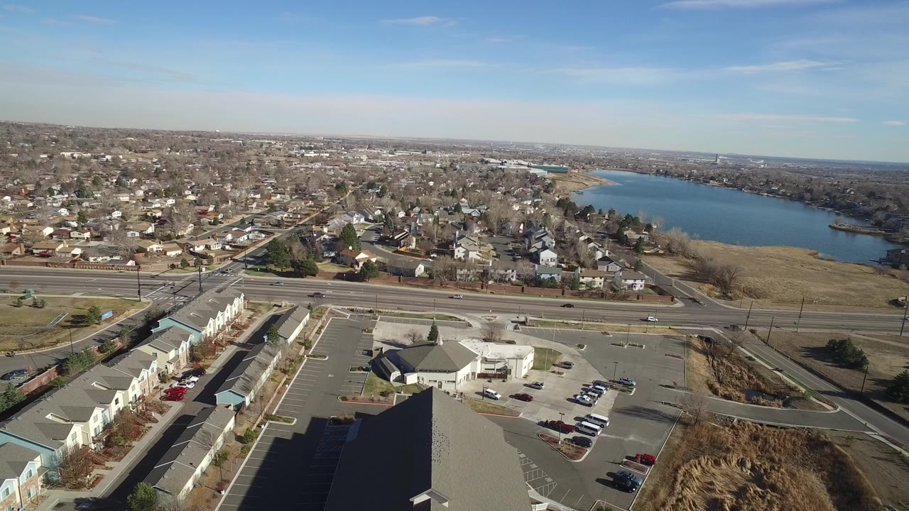Wide aerial frame over Hidden Lake, Colorado with a highway crossing the middle, homes beyond it, commercial roofs and parking lots below, blue lake water at right, and no visible people.