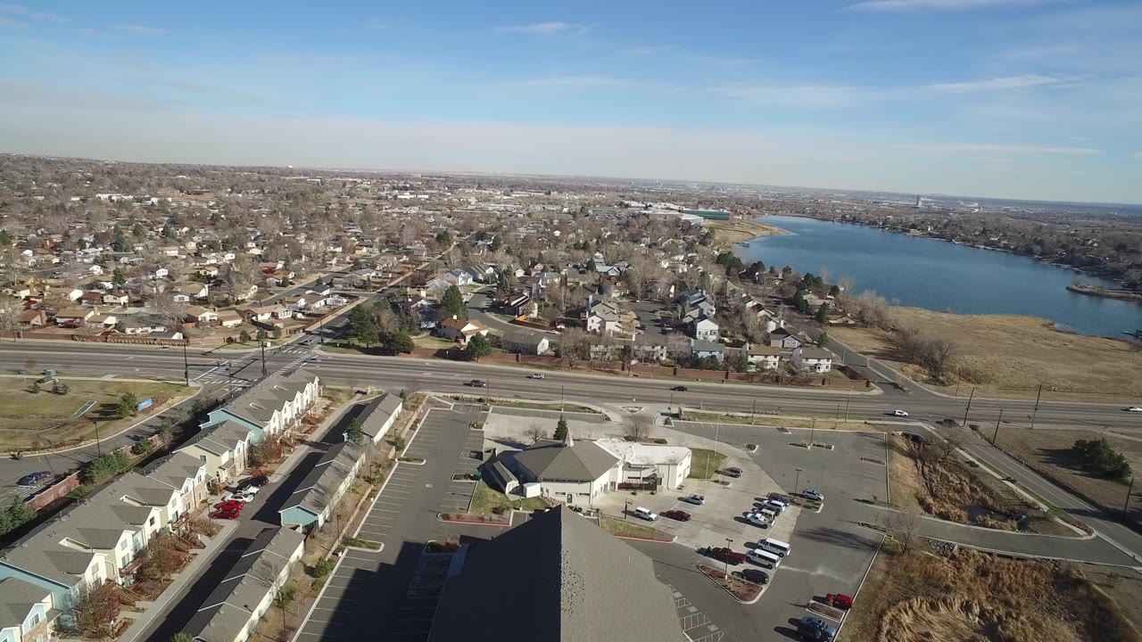 Aerial frame over Hidden Lake, Colorado with the highway running across the scene, a large parking lot and low buildings below, homes stretching across the background, lake shoreline at right, and no visible people.