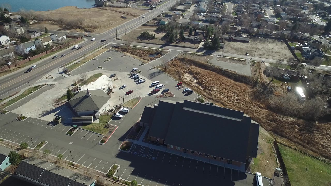 Aerial frame in Hidden Lake, Colorado with the dark-roofed building filling the lower frame, parking lots at left, a road across the upper left, and homes beyond open ground.